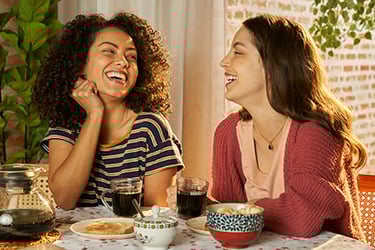 Duas mulheres sentadas em uma mesa com comidas e bebidas, tomando café da marca Café Damasco